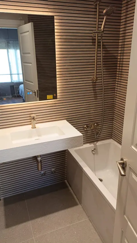 Baño con bañera, lavabo suspendido y pared de listones de madera con espejo retroiluminado